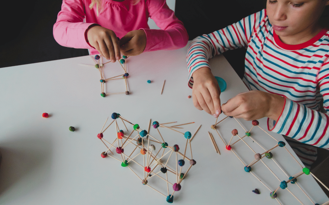 Two children build geometric structures from toothpicks and colorful beads on a white table during a craft STEAM for learners activity behind a striped shirted pair of kids.